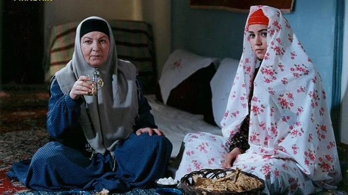 مینا جعفرزاده نشسته با روسری خاکستری در کنار بازیگری با چادر گل‌دار