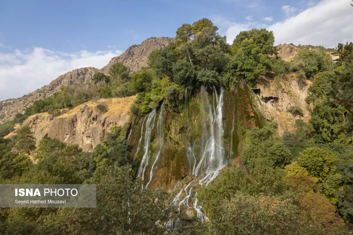 چشمانداز کامل آبشار بیشه نمای باز آبشار بیشه با دیواره سبز، صخرههای بالادست و آسمان آبی